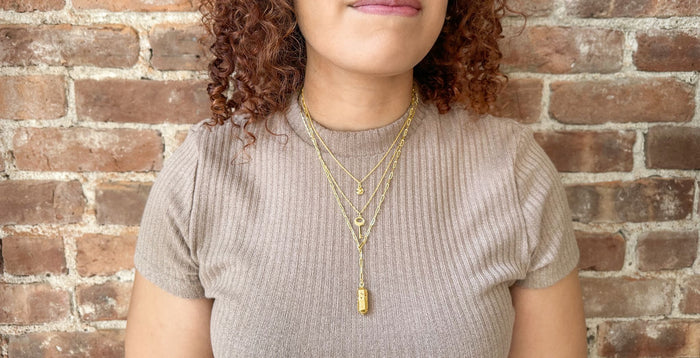A girl in front of a brick wall wearing a layer stack that consists of a gold Polygon Lariat Locket, Key Necklace (Medium), and a gold Mini Fortune Cookie Locket